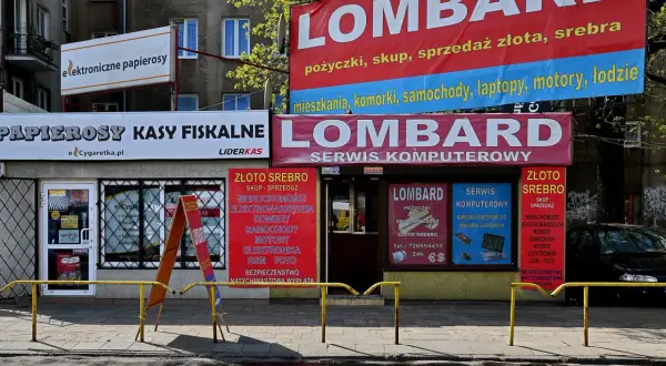 Szyldy lombardu i sklepów z elektroniką i papierosami na tle budynku mieszkalnego.